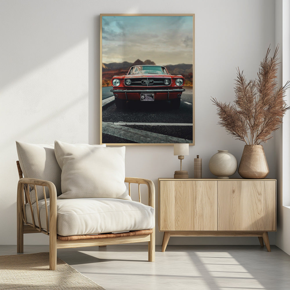 A low-angle, head-on shot of a vintage red Ford Mustang parked on an asphalt road. The iconic silver horse emblem is visible on the grille. In the background, a desert mountain range is blurred under a cloudy, muted sky. Poster