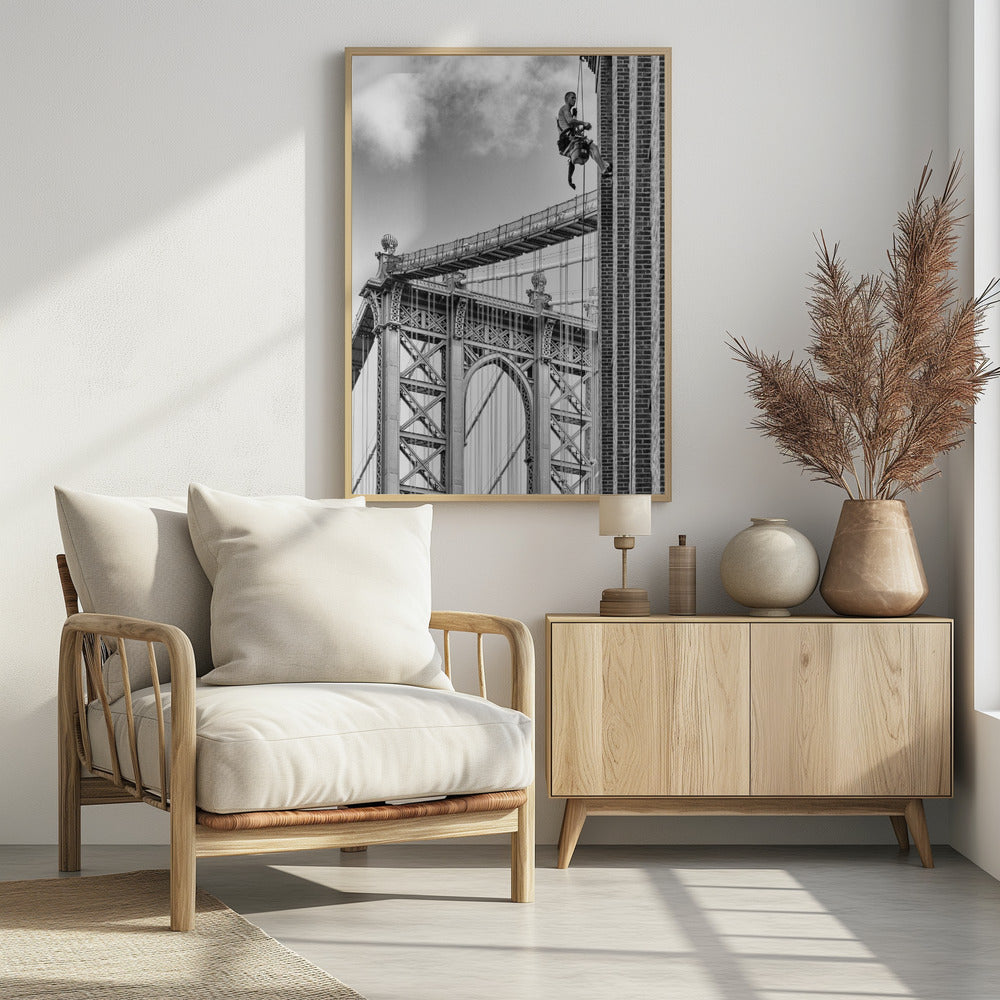 A black and white photograph capturing a window washer rappelling down the side of a tall brick building. In the background, the intricate steel structure of the Manhattan Bridge stands against a partly cloudy sky, creating a powerful urban scene. Decor