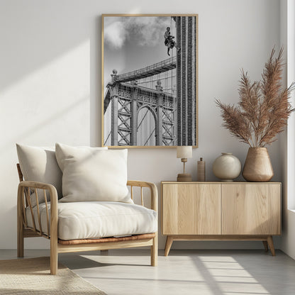 A black and white photograph capturing a window washer rappelling down the side of a tall brick building. In the background, the intricate steel structure of the Manhattan Bridge stands against a partly cloudy sky, creating a powerful urban scene. Decor
