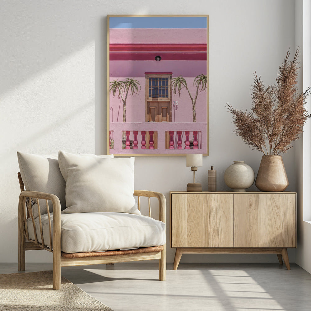 A brightly colored pink building stands under a clear blue sky, framed in black. A central wooden door with a paned window is flanked by two slender, palm-like trees. In the foreground, a pink balcony wall features fuchsia-colored balusters. Decor