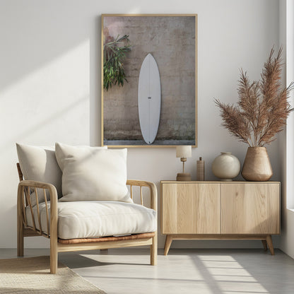A minimalist photograph of a white surfboard leaning against a textured beige concrete wall. To the left, a large green staghorn fern adds a touch of nature. The entire scene is enclosed in a simple black frame. Artwork