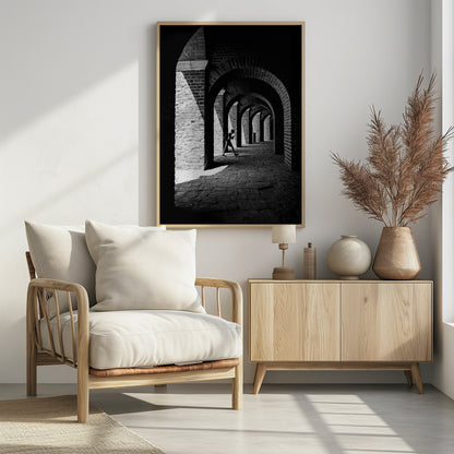 A dramatic black and white photograph of a person walking through a long brick arcade. Sunlight creates strong, contrasting shadows, highlighting the repeating arches that recede into the darkness, creating a powerful sense of depth and perspective. Decor