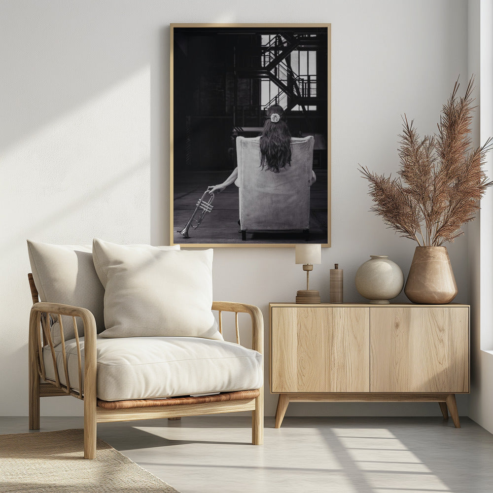 A black and white photograph of a woman seen from behind, sitting in a large, light-colored corduroy armchair. She has long, dark hair with a white flower pinned in it. Her left hand holds a trumpet, resting it on the dark floor of what appears to be an industrial warehouse with a metal staircase in the background. Wall Art