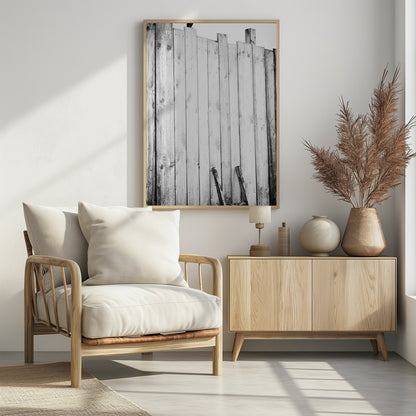 A black and white, low-angle photograph of a weathered wooden plank fence. The vertical planks are of varying heights, creating an uneven top line against a pale sky. The focus is on the texture of the wood, showing prominent grain, knots, and nail holes, all framed in black. Poster
