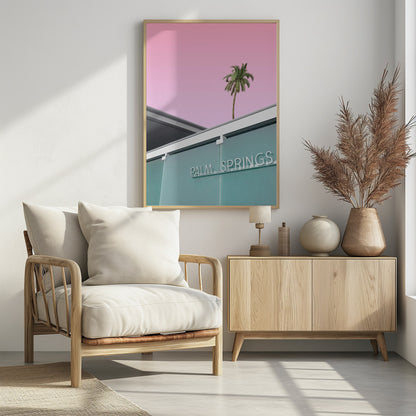 A low-angle shot of a modern building with a frosted teal glass facade and a white neon sign that reads 'PALM SPRINGS'. Above the building, a single tall palm tree stands against a solid, vibrant pink sky, creating a minimalist and stylized composition. Decor