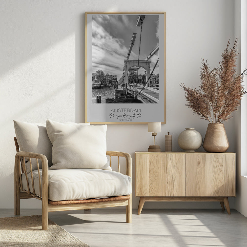 A black and white travel poster of Amsterdam, featuring a side view of the Magere Brug, a traditional wooden drawbridge, over the Amstel river. Classic canal houses line the waterway in the background under a dramatic, cloudy sky. The image is in a black frame and has text at the bottom that reads 'AMSTERDAM', 'Magere Brug, Amstel', and 'NEDERLAND'. Artwork