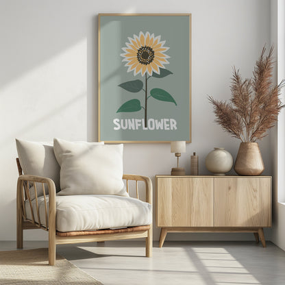 A stylized illustration of a single sunflower against a muted sage green background. The flower has yellow and white petals, a dark brown center, and a thin brown stem with green leaves. At the bottom, the word 'SUNFLOWER' is written in a bold, white, sans-serif font. The artwork is presented within a simple black frame. Decor