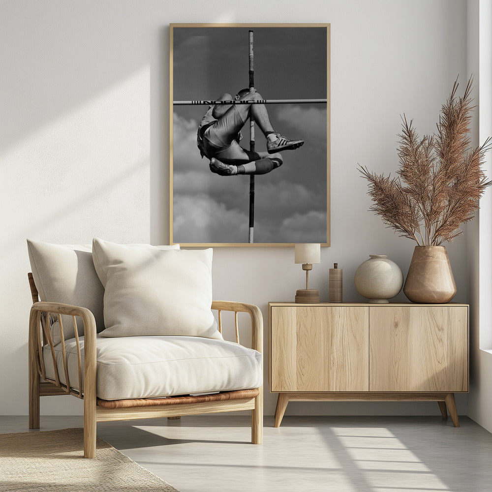 A dramatic black and white, low-angle photograph of a male athlete in mid-air during a pole vault. He is arching his back and bending his legs to clear the horizontal bar, with the vertical pole bisecting the frame. The background is a partly cloudy sky. Wall Art