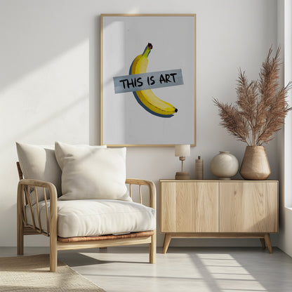 A framed artwork depicting a single yellow banana taped to a white wall with a grey strip of tape that reads 'THIS IS ART' in black marker-style font. Artwork