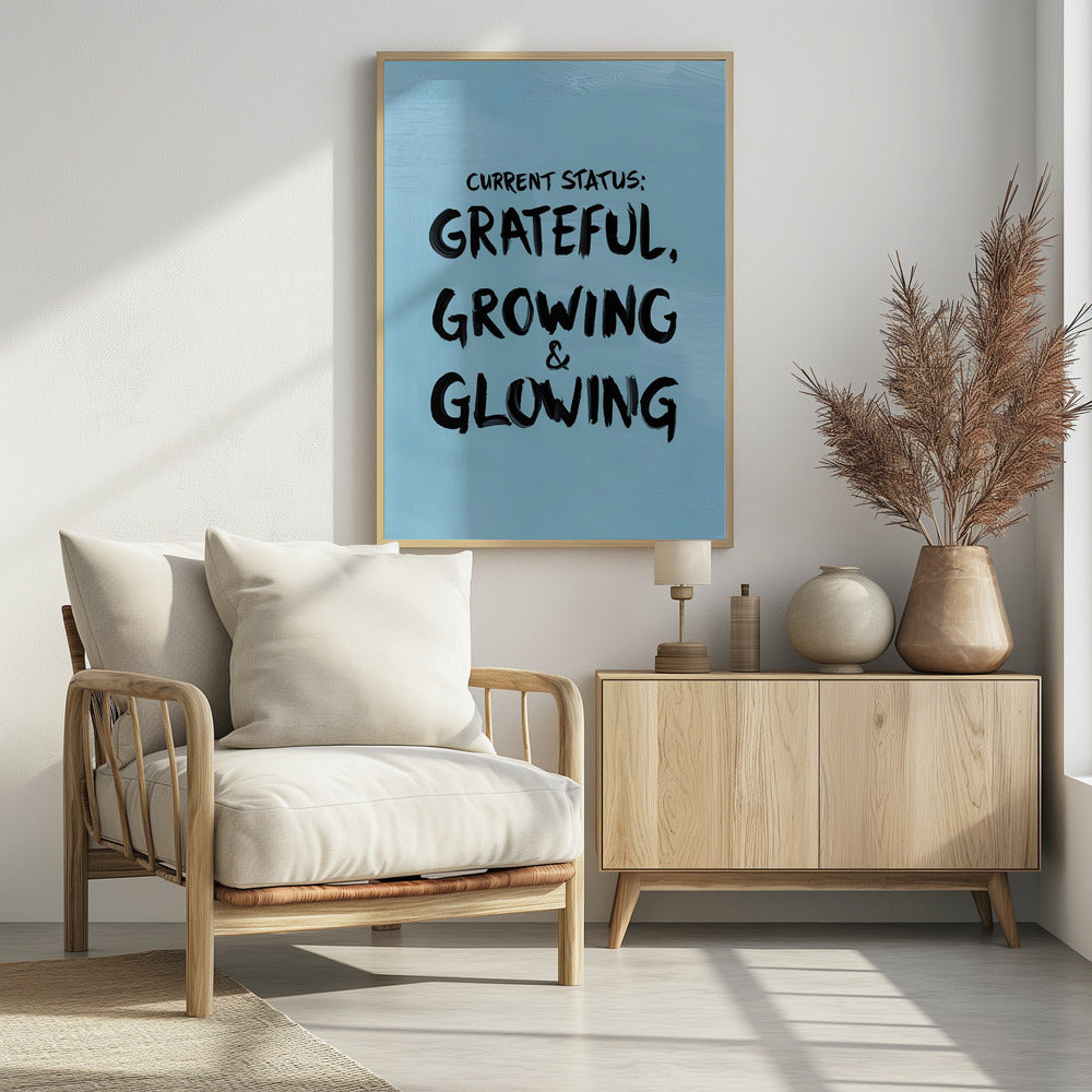 A motivational poster with a light blue background and a thin black frame. The text 'CURRENT STATUS: GRATEFUL, GROWING & GLOWING' is written in a bold, black, handwritten brush font. Decor