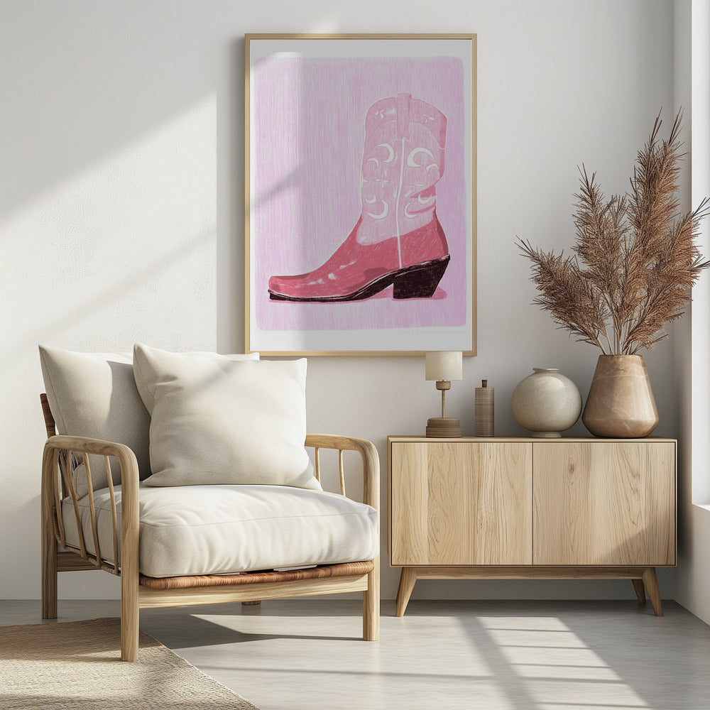 A framed art print of a pink cowboy boot against a light pink, textured background. The boot is illustrated in a block-print style with shades of pink, white decorative inlay patterns of moons and stars, and a dark brown heel and sole. Decor