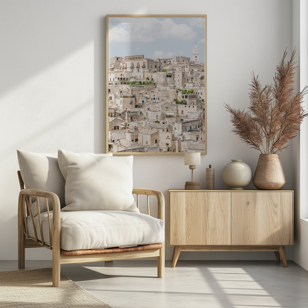 A vertical photograph of the ancient, stone city of Matera, Italy, with its dense collection of beige buildings and tiled roofs built up a hillside under a pale blue sky. A white bell tower stands out against the skyline. Poster