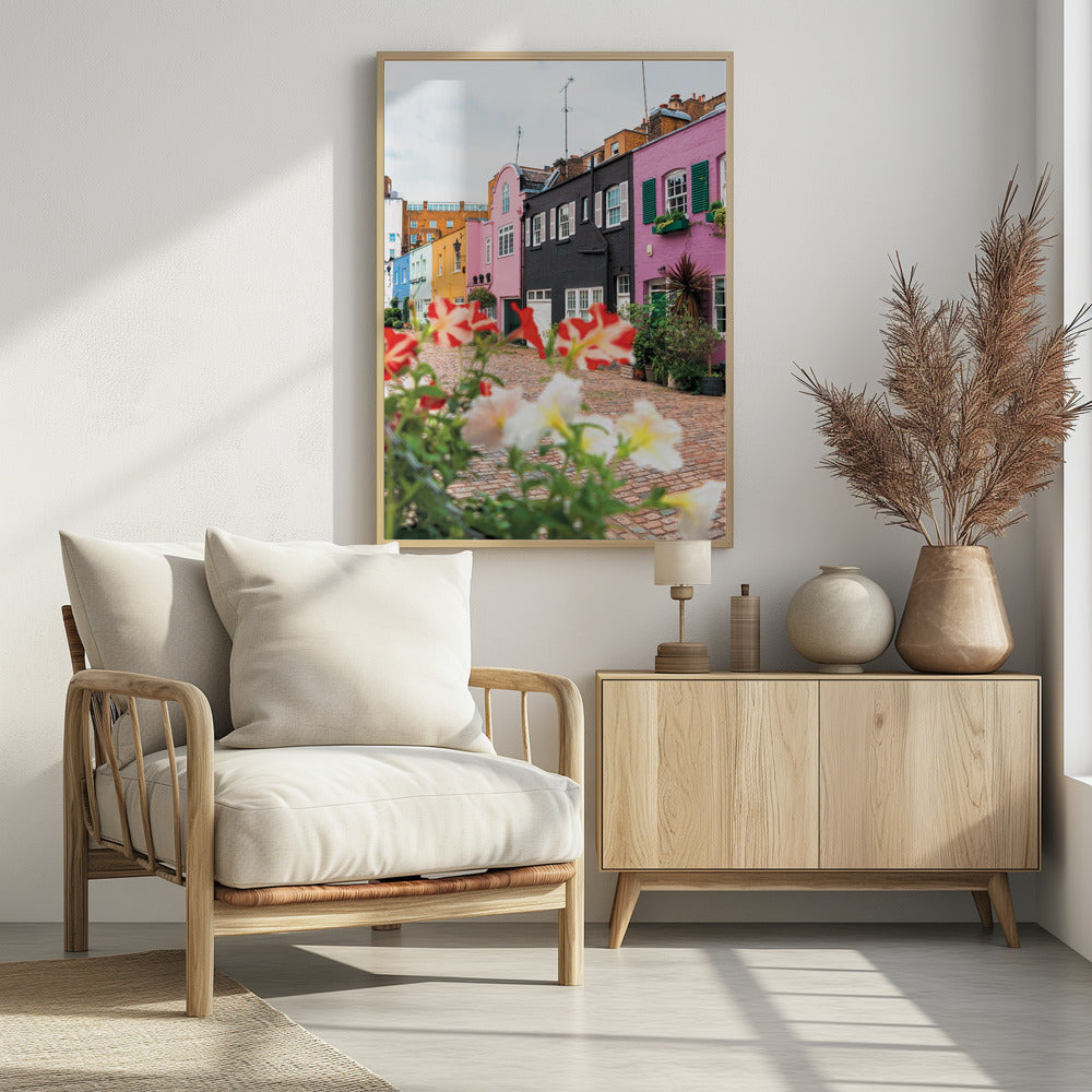 A low-angle view of a charming, narrow cobblestone street lined with colorful row houses in shades of pink, black, yellow, and blue, with blurred red and white petunias in the foreground. Wall Art