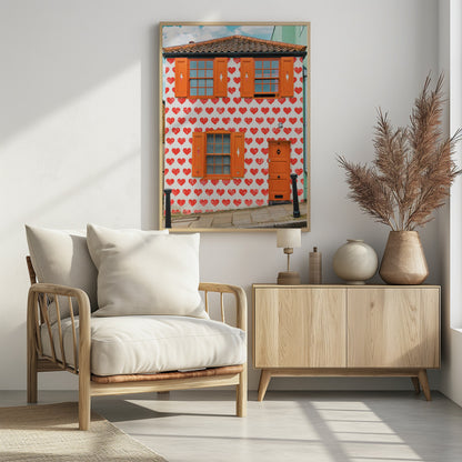 The front facade of a whimsical two-story house with white walls covered in a pattern of red hearts. The house has bright orange shutters on its windows and a matching orange front door, creating a vibrant and playful look against the heart-patterned background. Artwork