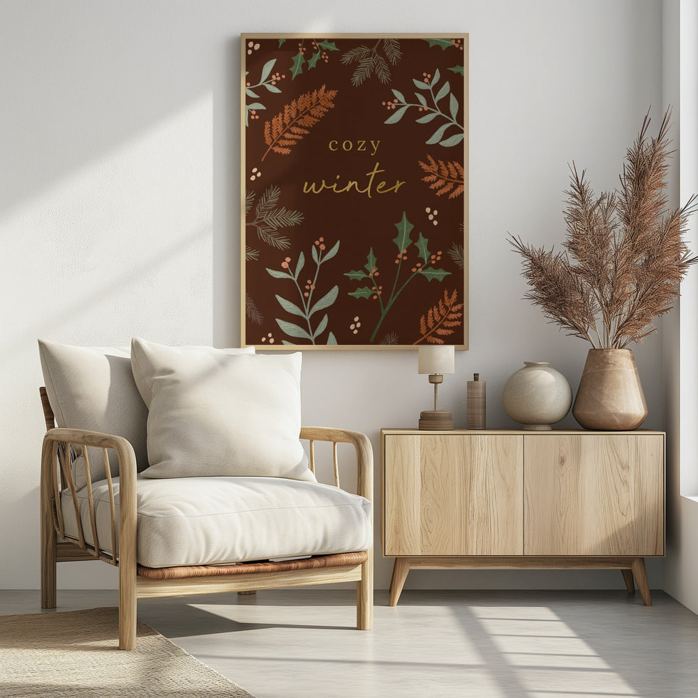 A warm, illustrative poster with a dark brown background featuring the words 'cozy winter' in gold script at the center. The text is framed by a wreath of winter botanicals, including holly, pine needles, and ferns in shades of green and burnt orange. Print