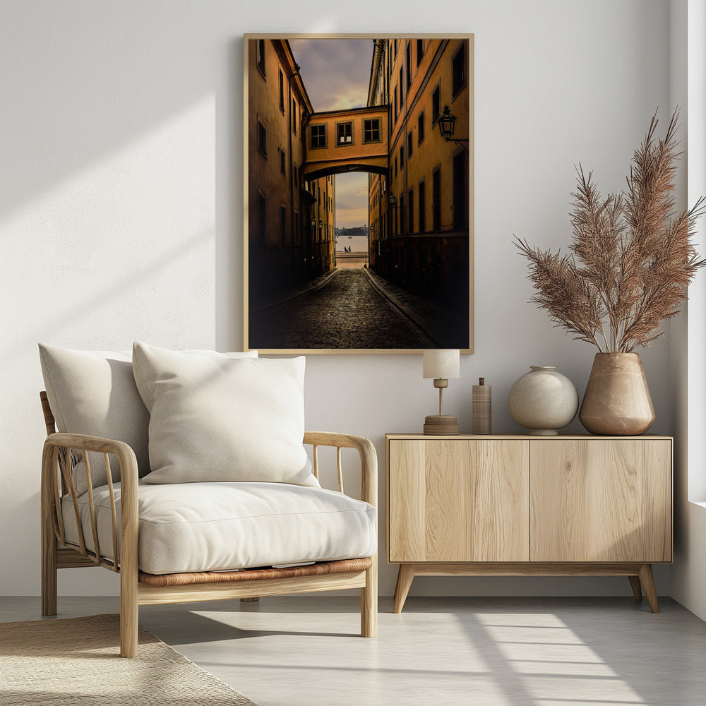 A perspective view down a narrow, shadowy cobblestone alley in an old European city, framed by warm, ochre-colored buildings. An arched walkway connects the buildings overhead, and the alley opens to a view of a tranquil body of water at sunset, with two small figures silhouetted by the shore. Decor
