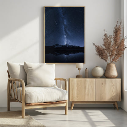 A breathtaking night photograph of the Milky Way galaxy stretching vertically across a deep blue, star-filled sky. Two shooting stars streak through the atmosphere above a dark, snow-dusted mountain range, which is perfectly reflected in the calm, mirror-like surface of a lake in the foreground. Artwork