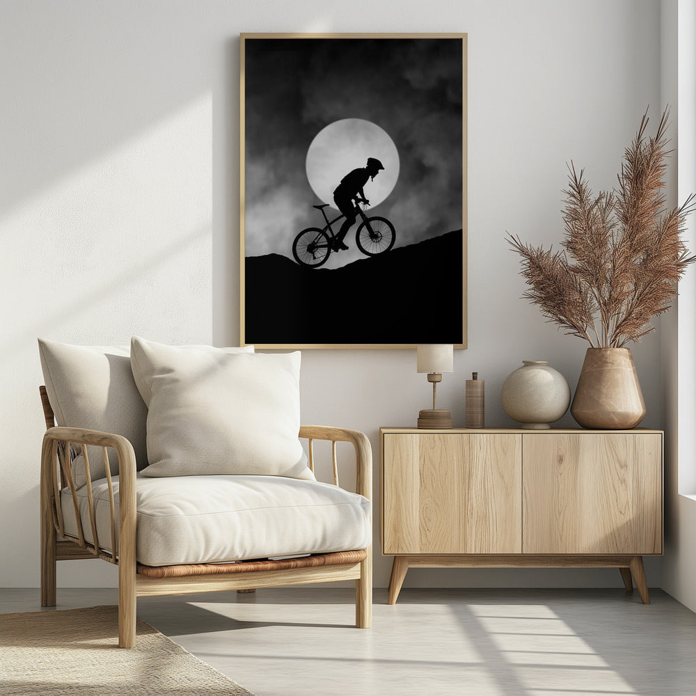 A dramatic black and white silhouette of a mountain biker riding uphill against the backdrop of a large, full moon in a cloudy night sky. Decor