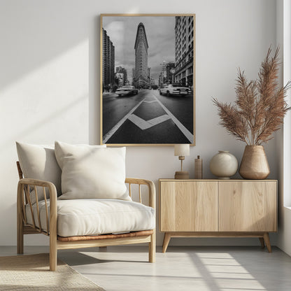 A dynamic black and white, low-angle photograph of the iconic Flatiron Building in New York City. The street in the foreground has bold white lines leading the eye to the building, while cars on either side are blurred with motion, capturing the city's fast pace under a cloudy sky. Decor