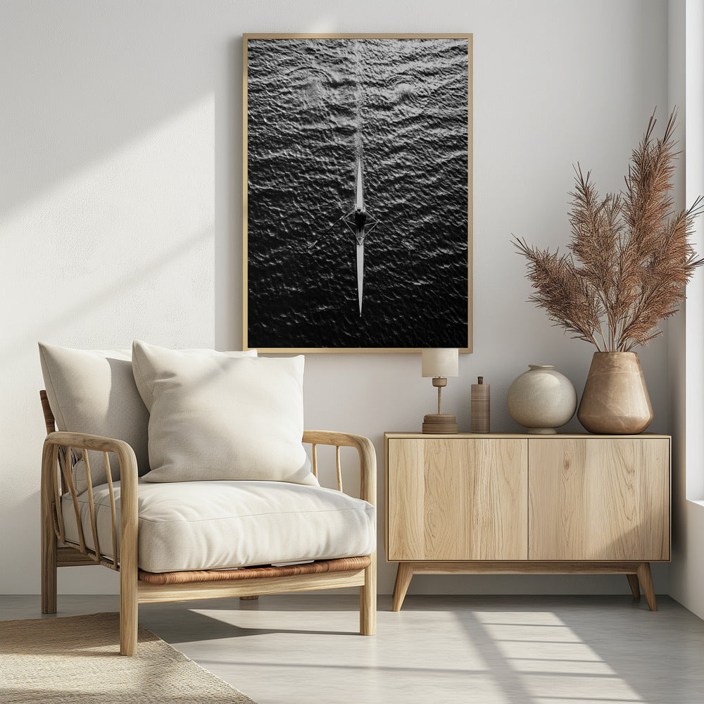 A dramatic black and white overhead photograph of a lone rower in a sleek racing shell, cutting a straight path through dark, textured water. The high-contrast lighting highlights the ripples on the water's surface and the rower's focused form. Artwork