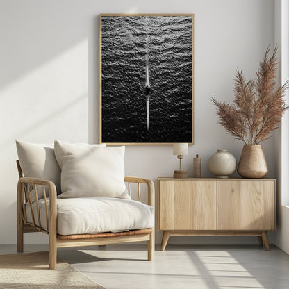 A dramatic black and white overhead photograph of a lone rower in a sleek racing shell, cutting a straight path through dark, textured water. The high-contrast lighting highlights the ripples on the water's surface and the rower's focused form. Artwork