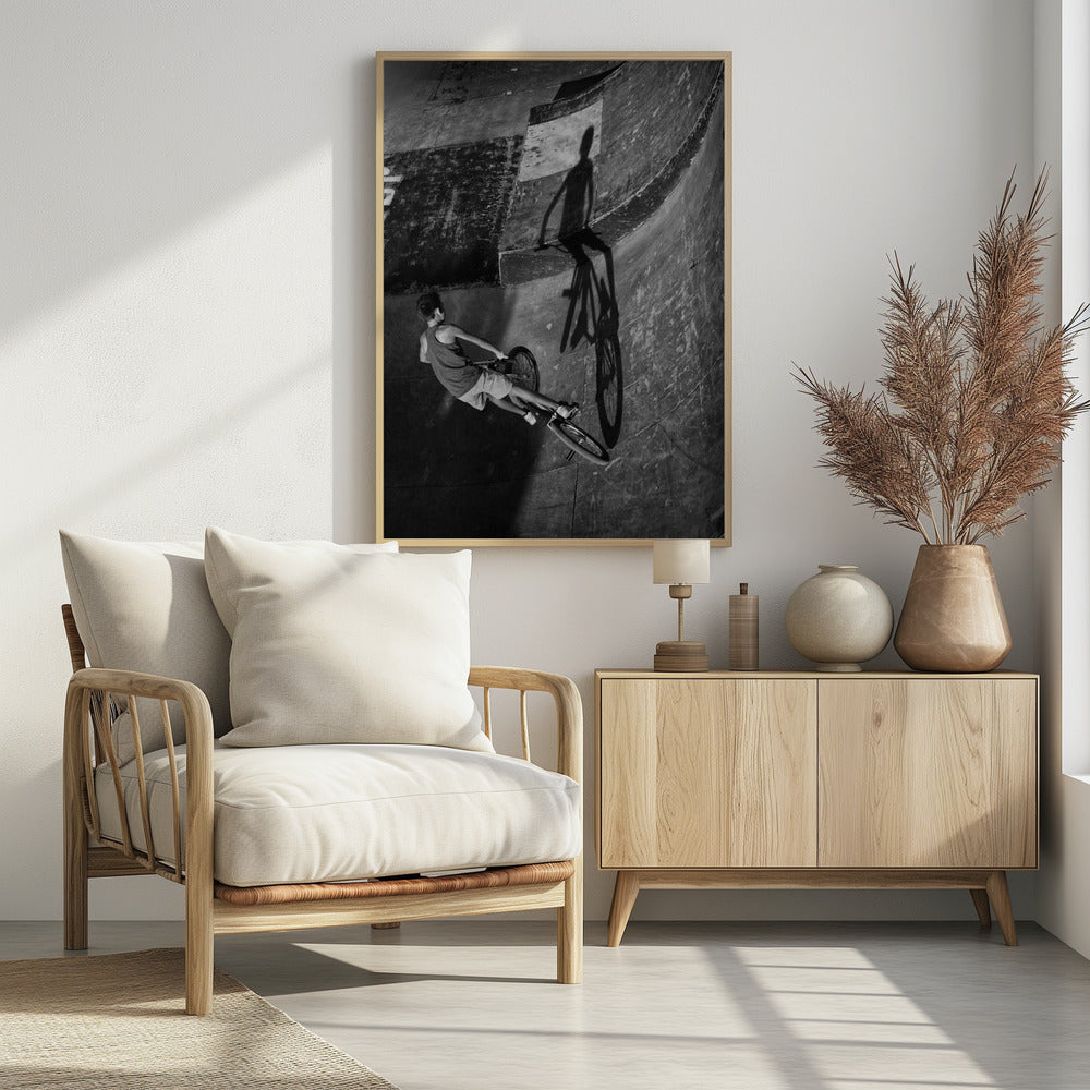 A dramatic black and white high-angle photograph of a BMX rider in a skate park. The rider is captured in motion on a curved ramp, while their long, stark shadow is cast on the worn, textured surface ahead, creating a powerful visual contrast. Poster