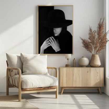 A mysterious black and white portrait of a woman wearing a large, black, wide-brimmed hat that casts a deep shadow over her eyes. Her hands are clasped at her chest, and the background is split vertically into black and white sections. Artwork