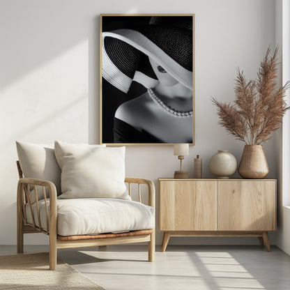A glamorous black and white close-up photo of a woman wearing a large, striped sun hat that hides her eyes, revealing only her dark lips. She wears a pearl necklace and an off-the-shoulder top against a dark background, creating a mysterious and elegant mood. Wall Art