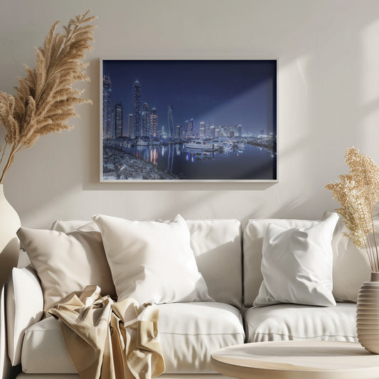 A stunning nighttime photograph of the Dubai Marina skyline, with illuminated skyscrapers towering against a deep blue sky. In the foreground, luxury yachts are moored in the calm harbor, their white hulls and the city's lights creating vibrant reflections on the water's surface. The shot is taken from a rocky embankment. Decor