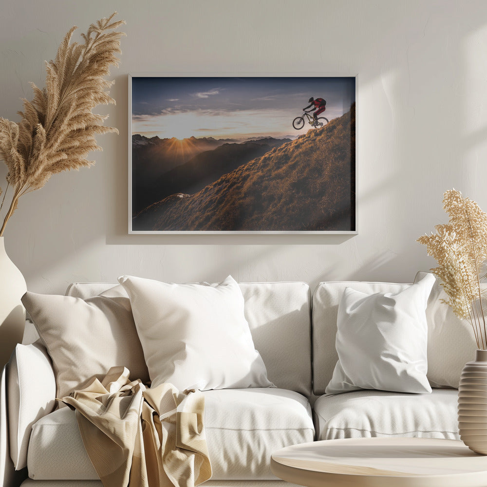 A mountain biker in mid-air, jumping down a steep grassy mountain ridge at sunrise. The sun is low on the horizon, casting a golden glow and long sunbeams over a vast, layered mountain range in the background. Poster