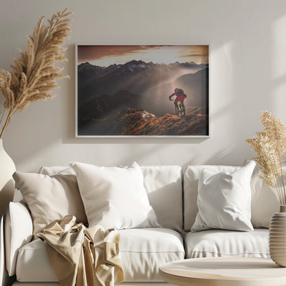 A mountain biker in red and black gear pauses on a grassy, sunlit ridgeline, looking out at a vast mountain range with snow-capped peaks as the sun sets behind them, casting dramatic rays of light through the valleys. Poster