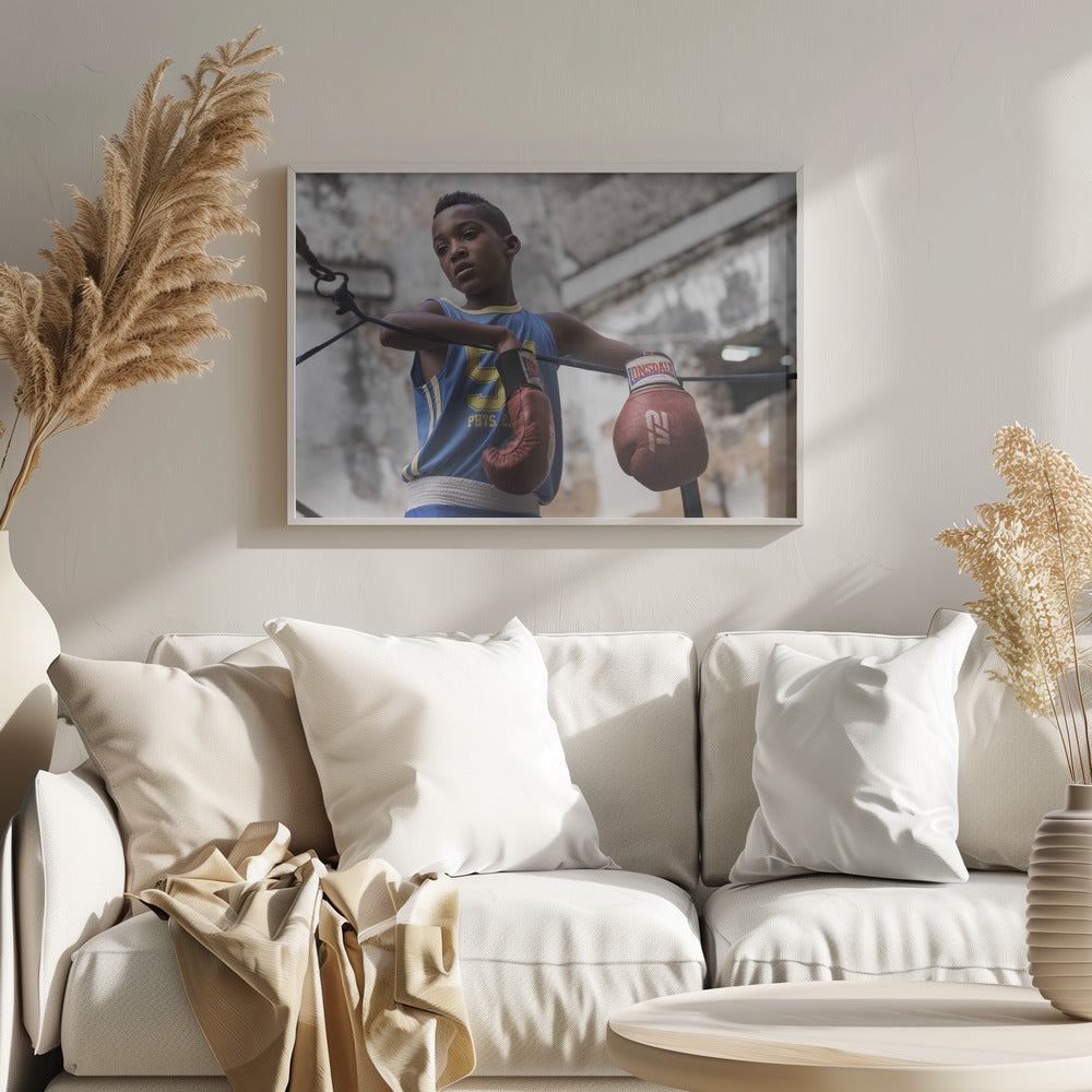 A young Black boy in a blue jersey and red boxing gloves leans pensively against the ropes of a boxing ring, looking away with a thoughtful expression against a blurred, weathered background. Wall Art