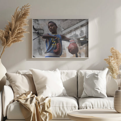 A young Black boy in a blue jersey and red boxing gloves leans pensively against the ropes of a boxing ring, looking away with a thoughtful expression against a blurred, weathered background. Wall Art