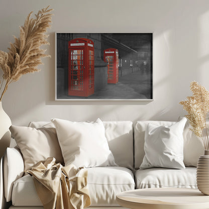 A selective color photograph of a city street at night, framed in silver. Two iconic red telephone booths are the only objects in color, standing out against the black and white background of the cobblestone sidewalk and iron fence. Artwork