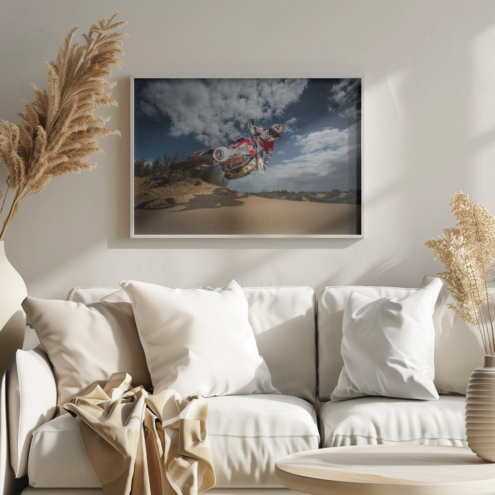 A low-angle action shot of a motocross rider on a dirt bike catching major air over a sand dune against a cloudy blue sky. The image is presented within a silver frame. Decor
