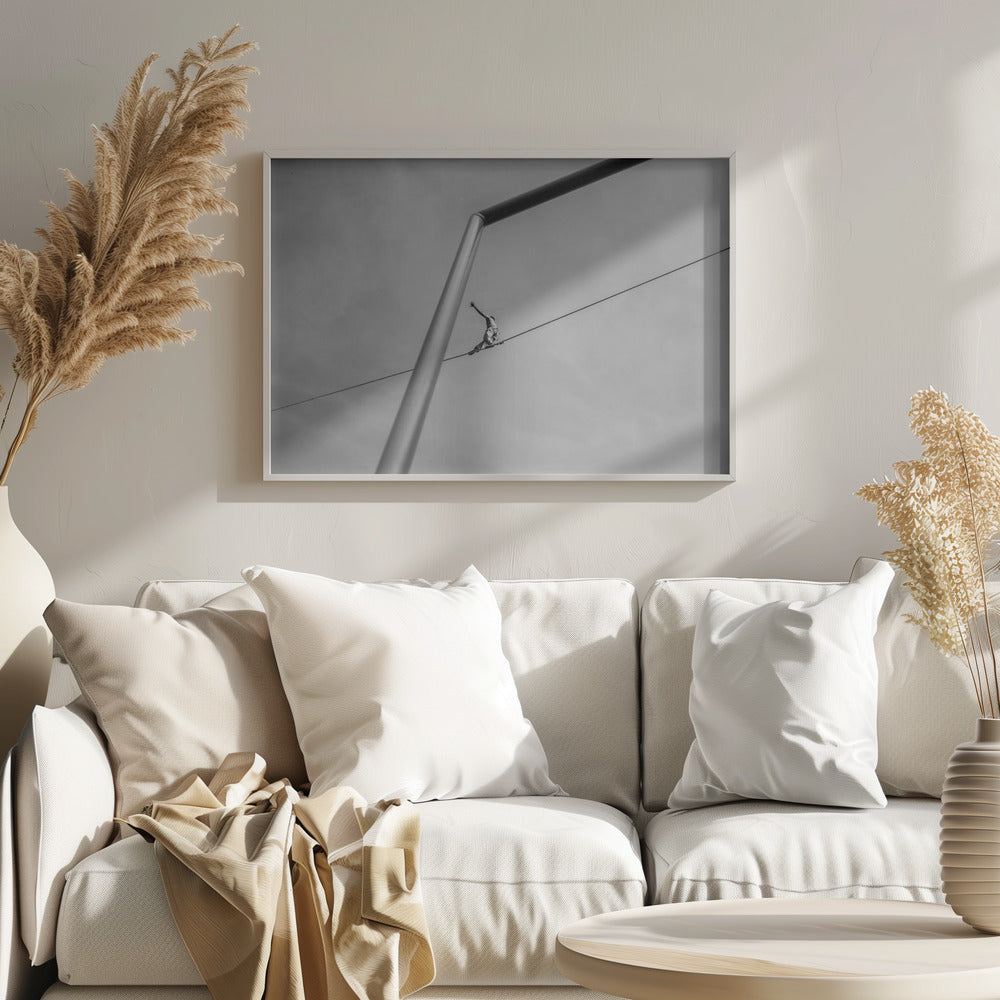 A black and white, low-angle photograph of a person balancing while sitting on a tightrope high in the sky. A large metal pole is in the foreground, creating a strong diagonal line. The image is framed in silver. Decor