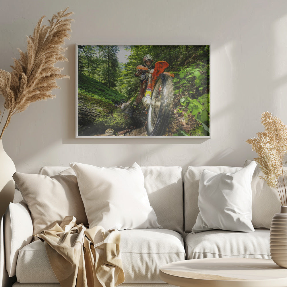 A motocross rider on an orange dirt bike tackles a muddy, wooded trail, maneuvering around a moss-covered log in a lush green forest, seen from a low-angle perspective. Decor