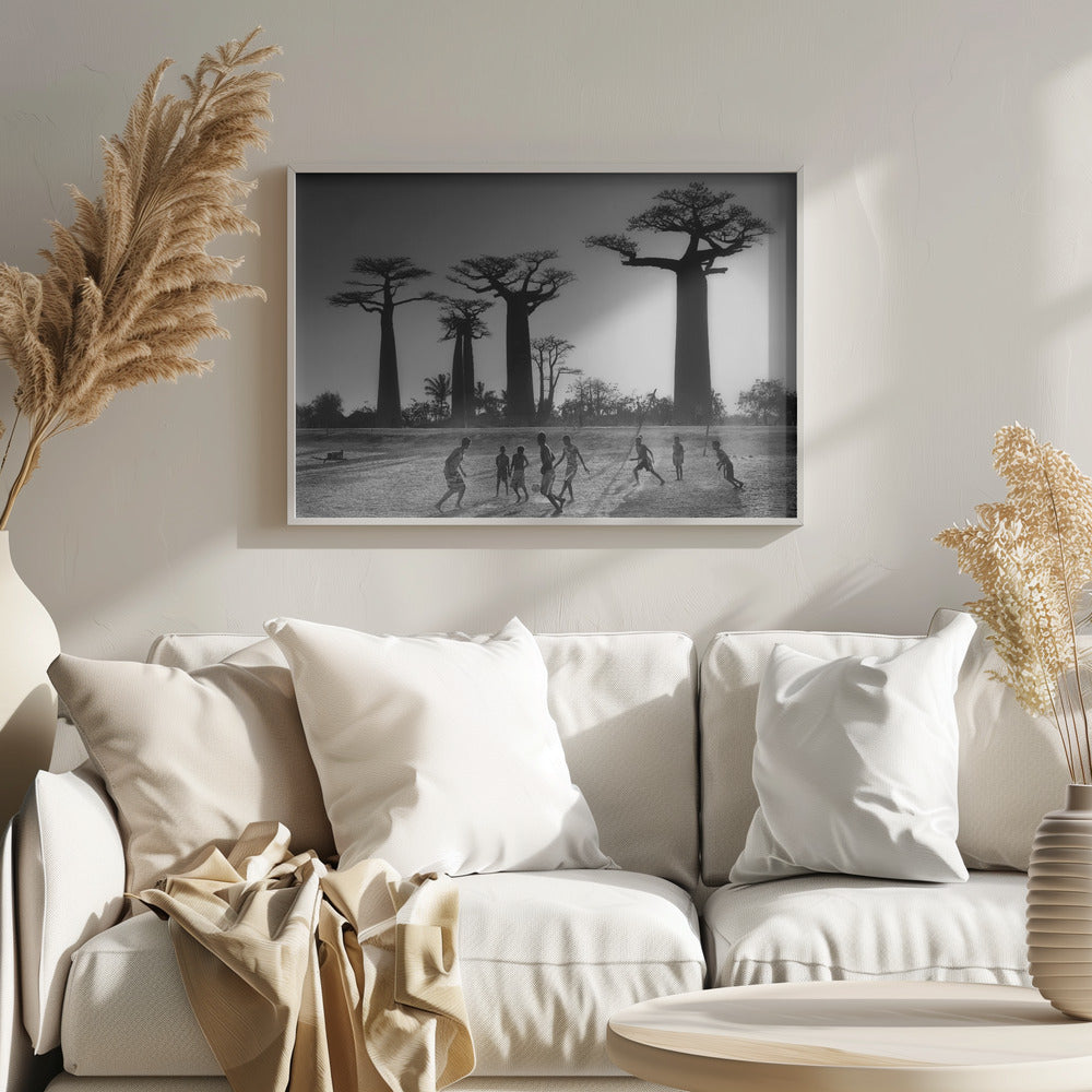 A framed, black and white photograph capturing a vibrant scene of several children playing soccer on a dirt field. In the background, the iconic silhouettes of large Baobab trees stand tall against a bright, hazy sun, casting long shadows of the players across the ground. Artwork