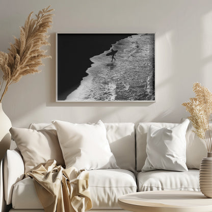 A high-contrast, black and white aerial photograph of a silhouetted surfer carrying a surfboard and walking along the shoreline. The image is divided diagonally, with the dark beach on the left and the white, foamy ocean waves on the right, creating a dramatic and graphic composition. The image is enclosed in a silver frame. Wall Art