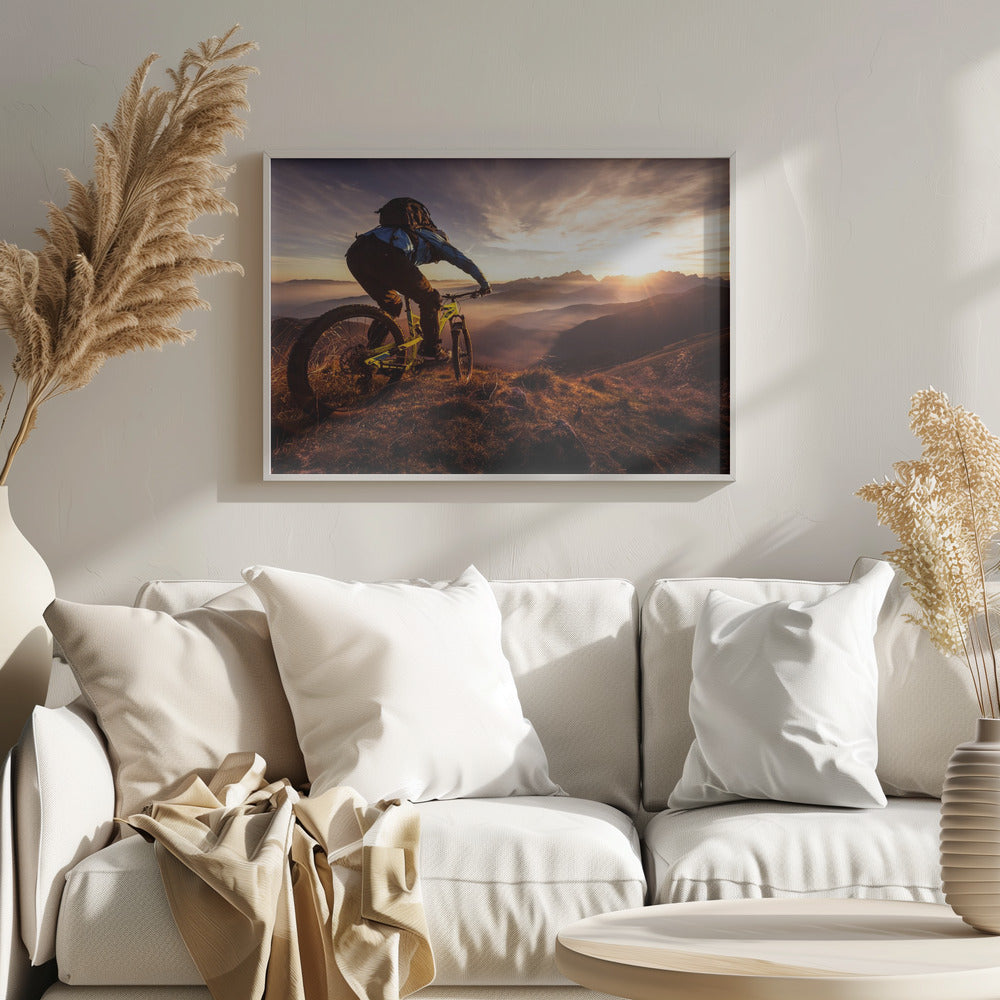 A mountain biker, seen from behind, stops at the top of a grassy hill to admire a spectacular sunset over a vast mountain range. The valleys below are filled with mist, and the sun's golden rays create a stunning starburst effect against the silhouetted peaks. Print