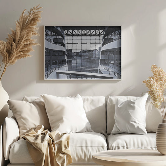 A symmetrical, black and white photograph taken from inside a modern building with curved balconies, looking out through a massive glass wall onto a canal and historic buildings on the opposite bank. A pedestrian bridge crosses the lower part of the frame. Decor