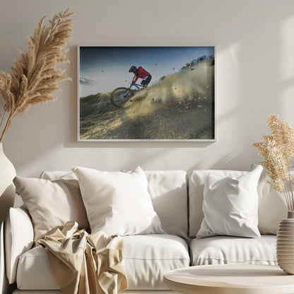 A mountain biker wearing a red jersey and helmet speeds down a steep, dusty trail, kicking up a large cloud of dirt. The sun is setting in the background, creating a lens flare against a clear blue sky. The image is captured from a low angle, emphasizing the action and speed, and is presented within a silver frame. Artwork