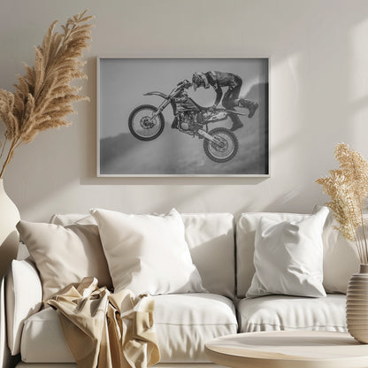 A dynamic black and white photograph of a motocross rider in mid-air, performing a jump. The rider, wearing full protective gear and a helmet, is leaning forward over the handlebars of the dirt bike. The bike is suspended against a blurry, light gray background, emphasizing the action and height of the jump. The image is enclosed in a silver frame. Wall Art