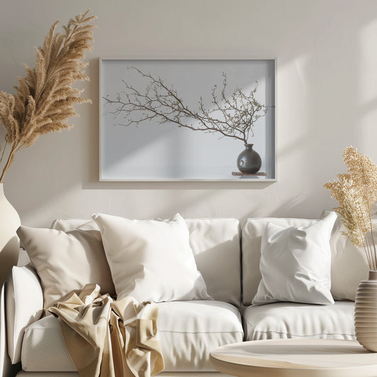 A minimalist still life of a long, delicate branch with small leaves arranged horizontally in a small, dark, round ceramic vase. The background is stark white, creating a high-contrast, serene composition, all within a silver frame. Print