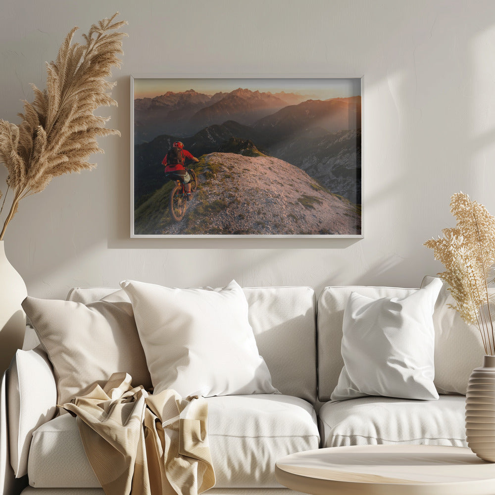 A mountain biker wearing a red shirt and helmet rides along a narrow, rocky mountain ridge at sunset. The background features a vast landscape of layered mountain ranges glowing in the warm, golden light of the setting sun. Poster