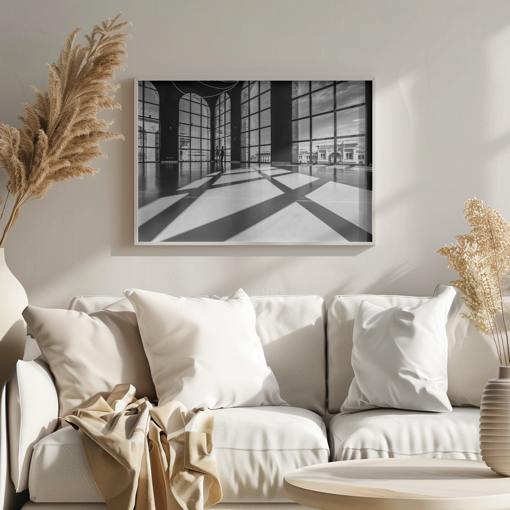 A striking black and white photograph of a spacious, modern hall with a polished floor. Dramatic shadows from large, arched windows are cast across the floor. Through the windows, a view of a classical European city with a triumphal arch can be seen. Print