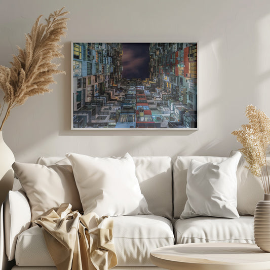 A low-angle nighttime view looking up from a courtyard surrounded by extremely dense, stacked apartment buildings with many windows lit in yellow, blue, and white, revealing a small patch of dark, cloudy sky. The image is presented within a brushed silver frame. Artwork