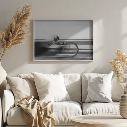 A black and white photograph captured with motion blur, showing a cyclist in an aerodynamic position racing at high speed. The background is blurred into horizontal lines, emphasizing the velocity. The image is enclosed in a silver frame. Wall Art