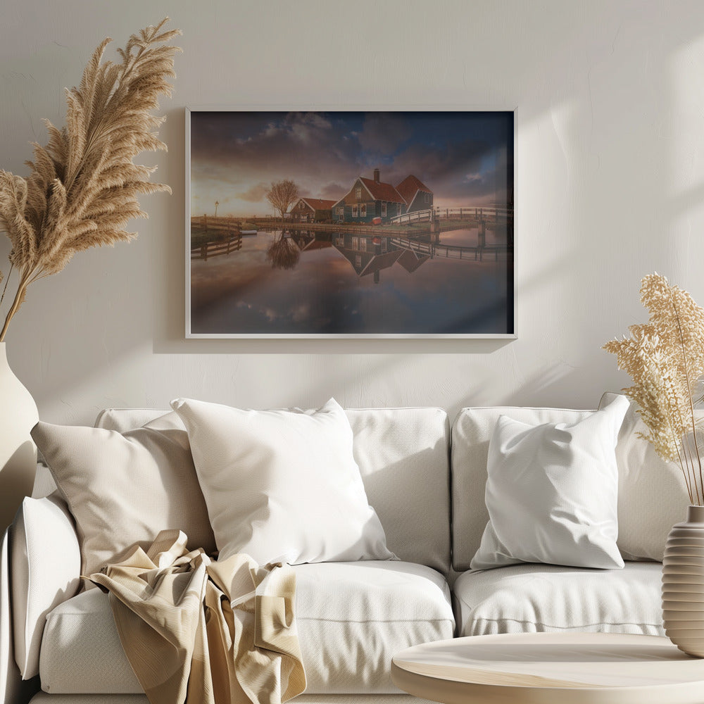 A picturesque landscape of traditional Dutch houses with teal walls and red roofs reflected perfectly in the calm water of a canal at sunset, under a dramatic, cloudy sky. A small wooden bridge crosses the canal, and the scene is enclosed in a silver frame. Artwork