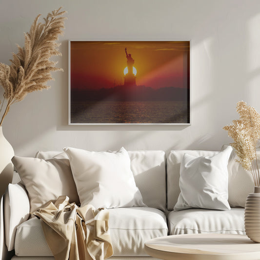 A powerful silhouette of the Statue of Liberty against a fiery orange and red sunset. The sun is perfectly positioned behind the statue's base, creating a brilliant, glowing orb of light. The dark waters in the foreground reflect the vibrant colors of the sky. The photograph is presented within a silver-colored frame. Artwork